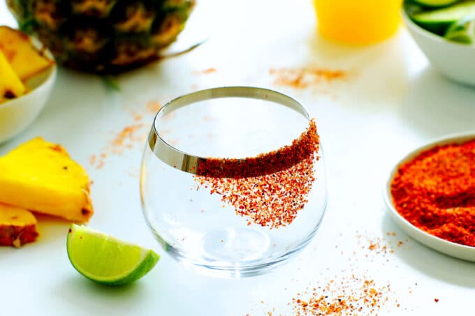 an empty glass with tajin on one side, pineapple, lime wedges, and a plate of tajin in the background