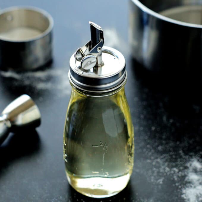 A glass bottle of simple syrup with a pour spout lid next to a sauce pan and measuring cups