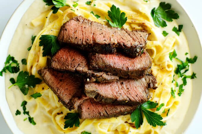 An overhead shot of slices of steak laying atop a plate of noodles and Alfredo sauce with a parsley garnish