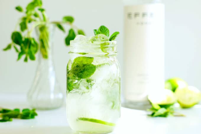 A vodka mojito in a mason jar with mint and lime garnish and a bottle of vodka in the background