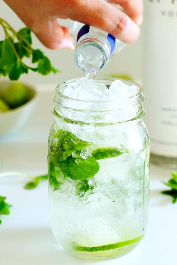 Vodka being poured into a mason jar with ice, mint, and lime in it