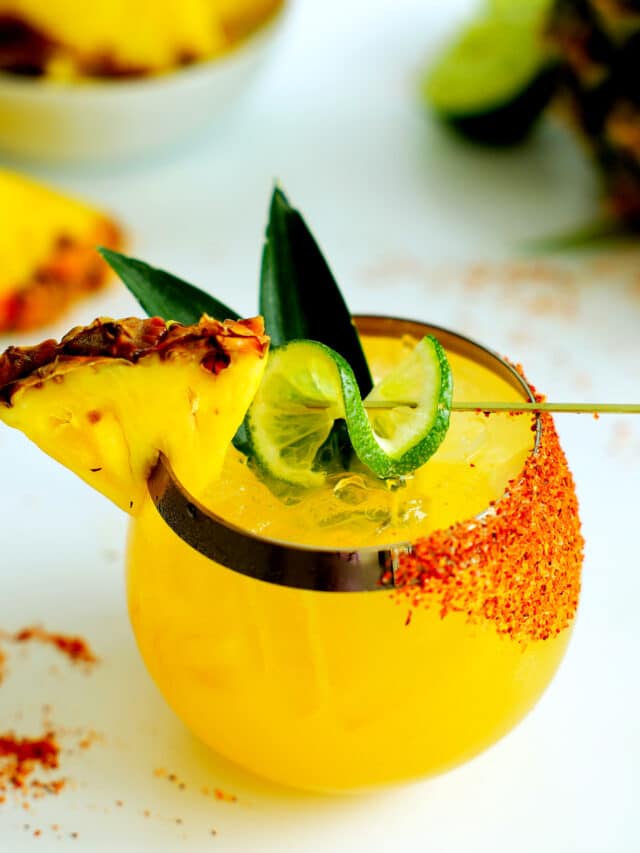 A pineapple margarita with a lime twist, pineapple wedge, and pineapple leaves garnish and tajin on one side of the glass