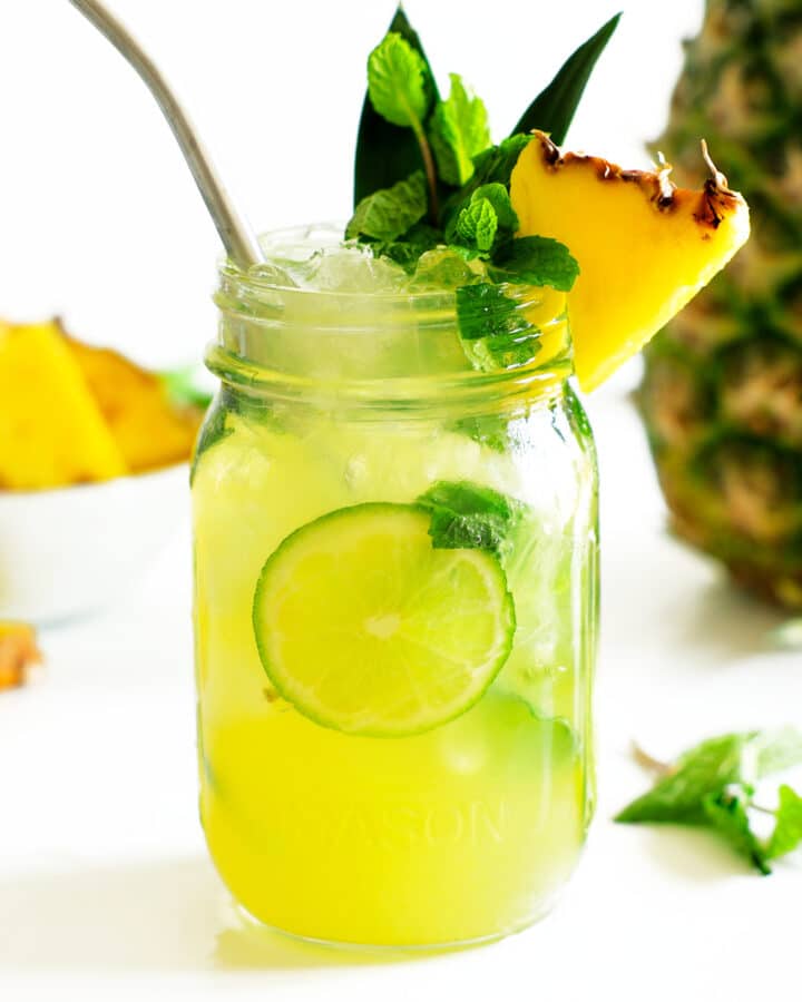 A pineapple mojito in a mason jar with pineapple and mint garnish and pineapple in the background