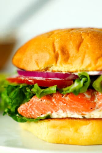 A a close up side view of a fully assembled salmon sandwich with red onions, tomatoes, and lettuce