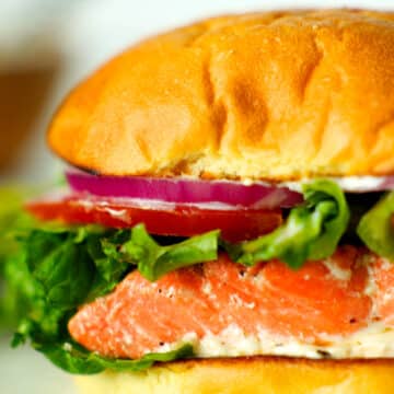 A a close up side view of a fully assembled salmon sandwich with red onions, tomatoes, and lettuce