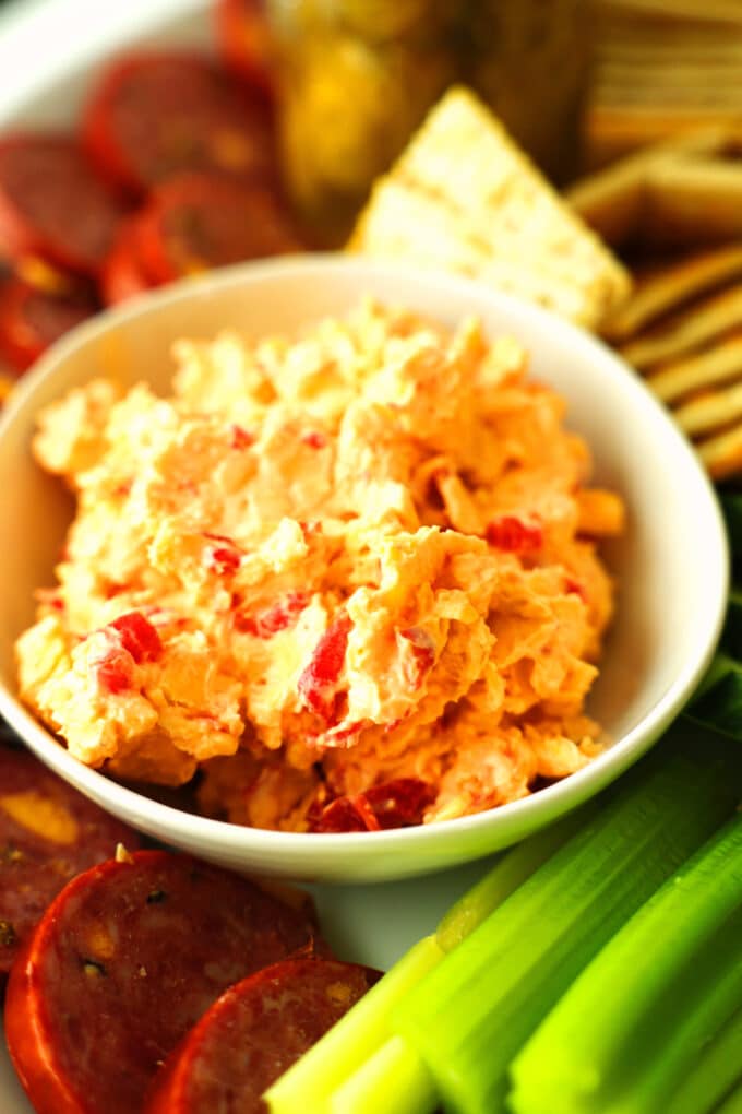a close up of a bowl of pimento cheese with celery, summer sausage, and crackers around it