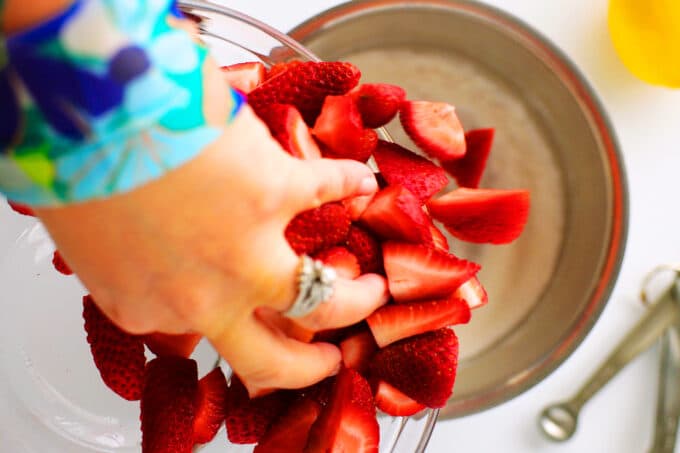 A hand scooping sliced strawberries from a bowl into a saucepan