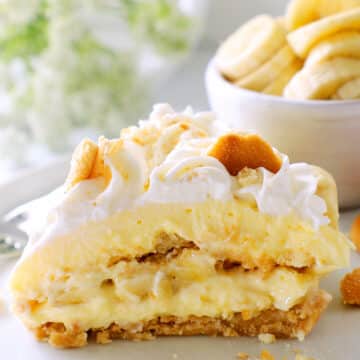 a side view of a slice of banana pudding cheesecake showing the different layers. a bowl of banana slices in the background