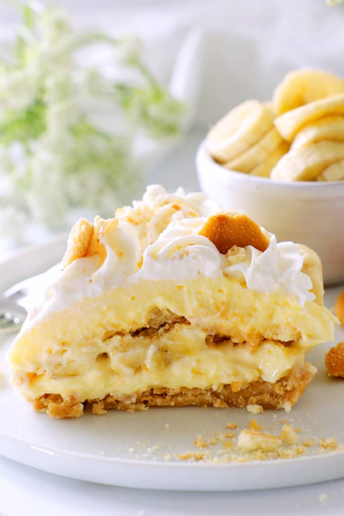 a side view of a slice of banana pudding cheesecake showing the different layers. a bowl of banana slices in the background