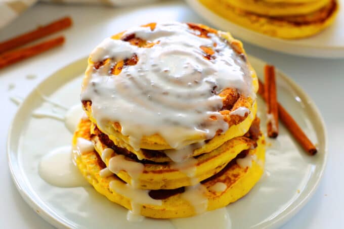 a stack of cinnamon roll pancakes on a plate with vanilla glaze drizzled over top and cinnamon sticks beside it