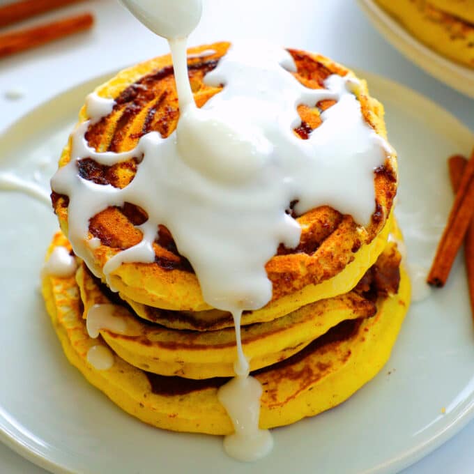 a stack of cinnamon roll pancakes on a plate with vanilla glaze drizzled over top and cinnamon sticks beside it