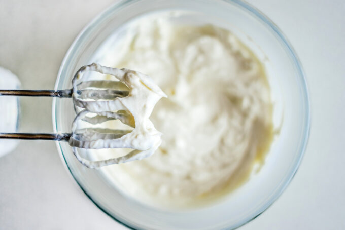 the beaters of a hand mixer with stiff peaks of whipped cream on them and a bowl of whipped cream below