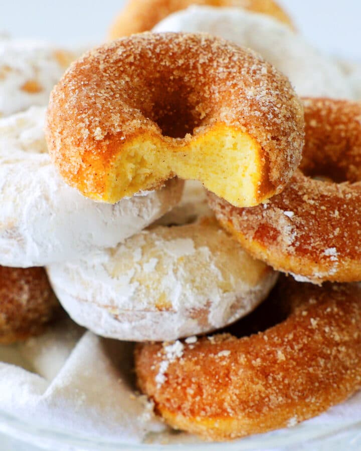 A stack of cinnamon sugar and powdered sugar donuts with a bite taken out of the top one. the inside is yellow and dense.