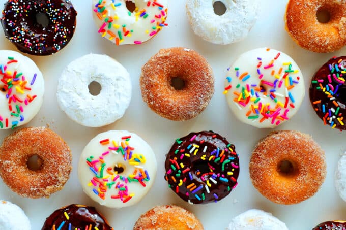 An overhead view of donuts layer out in rows with all different toppings. Chocolate with sprinkles, vanilla with sprinkles, cinnamon sugar, and powdered sugar.