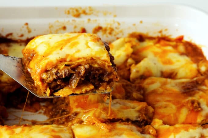 A spatula lifting a serving of beef enchilada casserole out of the dish. The layers of cheese, ground beef, and tortillas are visible.