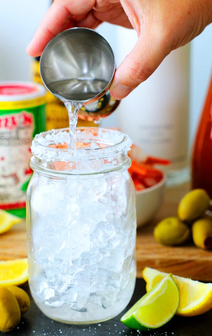 vodka being poured from a jigger into a jar of ice with a salt rim