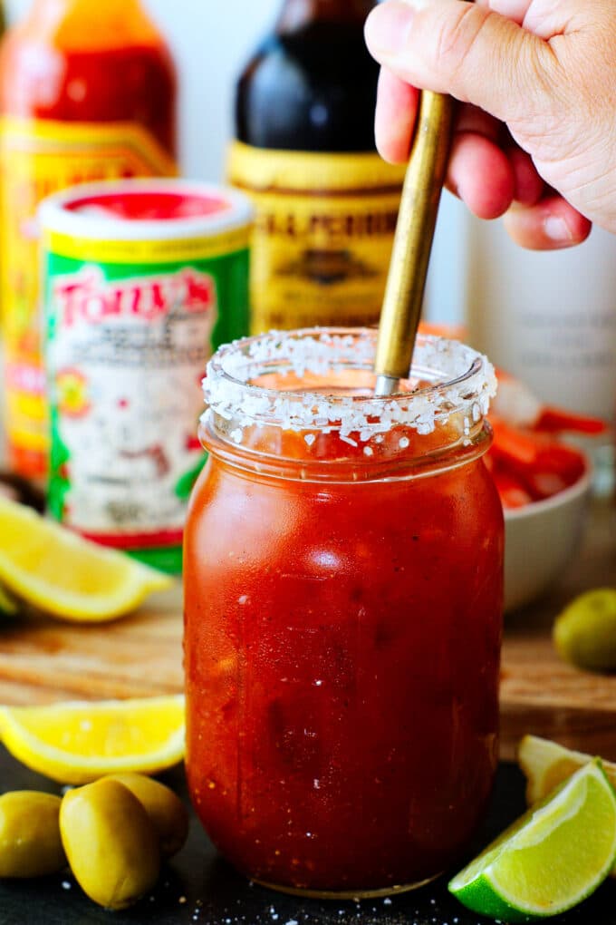 A hand stirring a Bloody Mary in a jar, with a salt rim, ingredients in the background