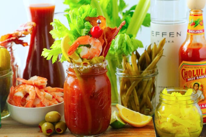 A Bloody Mary in a jar with a salt rim. the mouth of the glass is stuffed with garnishes, bacon, shrimp, lemon wedge, olives, and celery sticks. more of the garnishes sit around it in bowls and jars with hot sauce and vodka in the background
