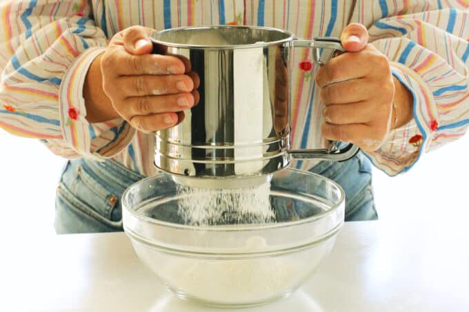 a woman sifting dry ingredients into a bowl with a sifter