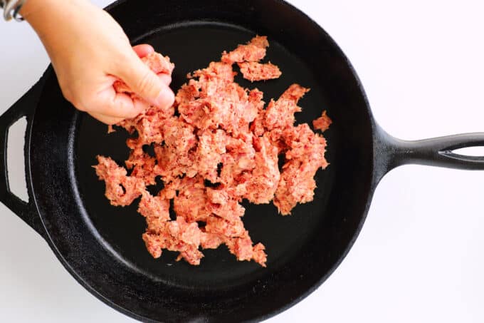 a hand breaking up raw breakfast sausage and adding it to a cast iron pan