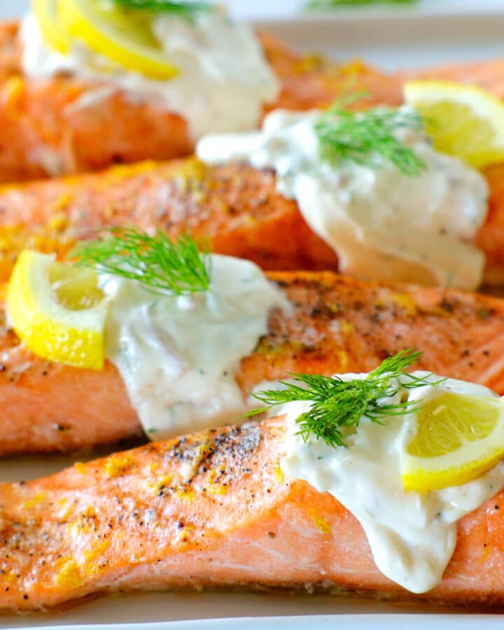A side angle of a row of salmon fillets, creamy, white dill sauce falling over the sides and fresh dill and lemon slices for garnish
