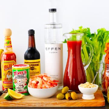 A horizontal shot of all of the Bloody Mary Bar ingredients neatly arranged on a wooden board against a white wall