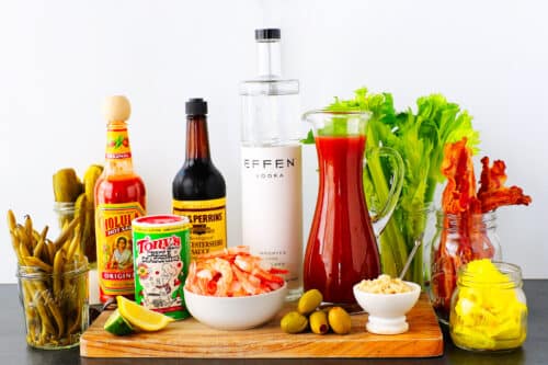 A horizontal shot of all of the Bloody Mary Bar ingredients neatly arranged on a wooden board against a white wall