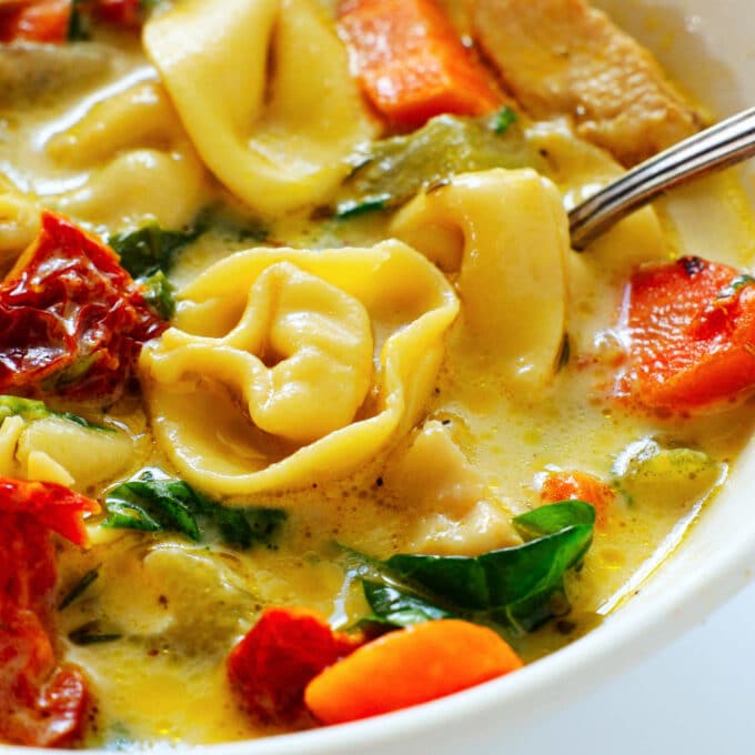 A close up of Chicken Tortellini Soup in a bowl. Noodles, sun dried tomatoes, spinach, and carrots are all visible in the creamy broth