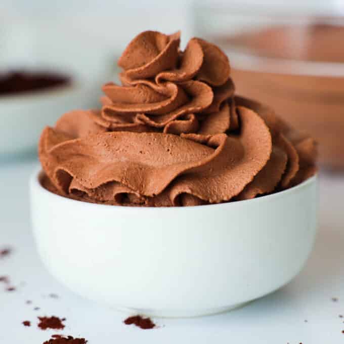 Chocolate Whipped Cream piped into a small, white bowl. a bit of cocoa powder is spilled on the counter beside it.