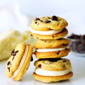 A stack of three chocolate chip sandwich cookies with a fourth leaning against them, a bowl of chocolate chips in the background