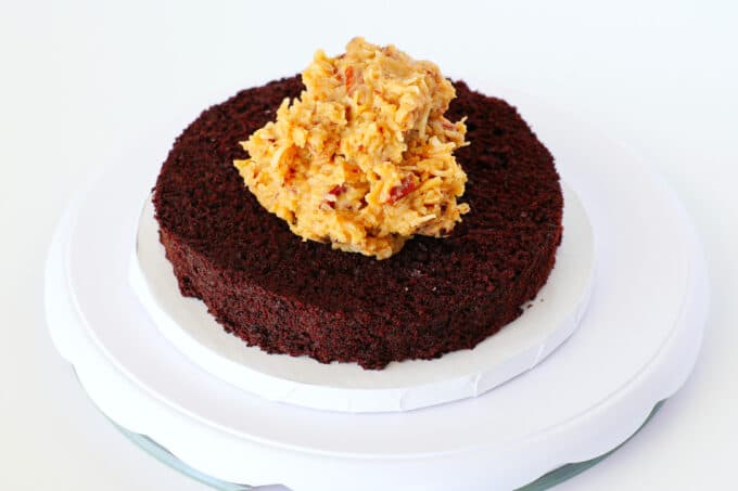 A big blob of German chocolate cake frosting sits on top of the first cake layer on a cake plate