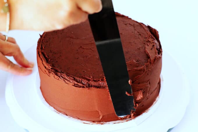 the final layer of chocolate buttercream frosting being added to the sides of the cake with an icing spatula