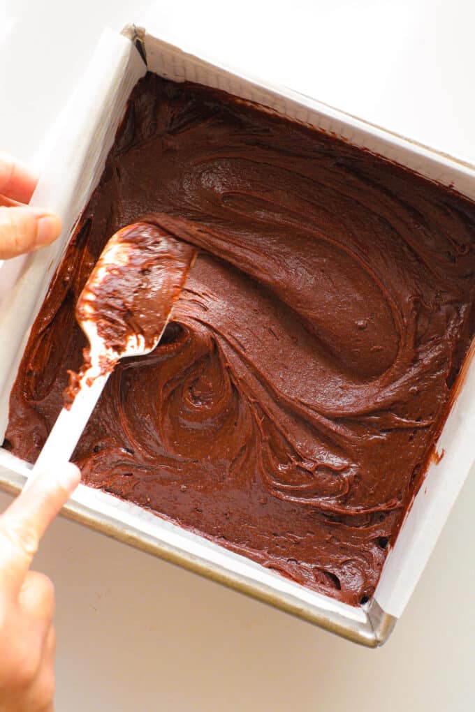 the brownie batter being spread into a square pan with a silicon spatula