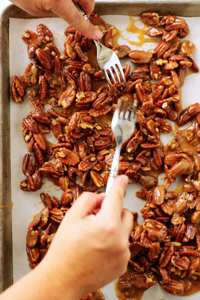 an overhead view of a persons hands using two forks to separate the praline pecans on a sheet of parchment paper