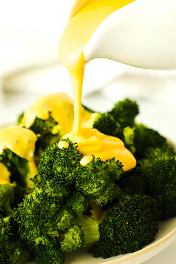 creamy yellow cheese sauce being poured over a plate of steamed broccoli florets