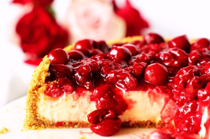 A close up of a cherry cheesecake that has been cut into. The layers of crust, filling, and cherry topping are all visible from the side