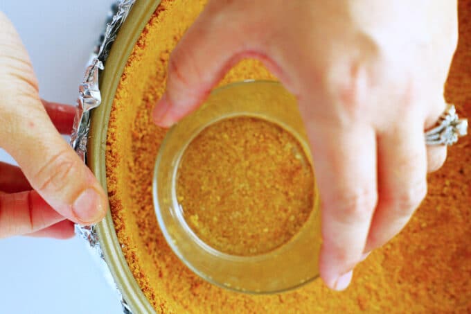 hands using a glass to press the graham cracker crust into the pan