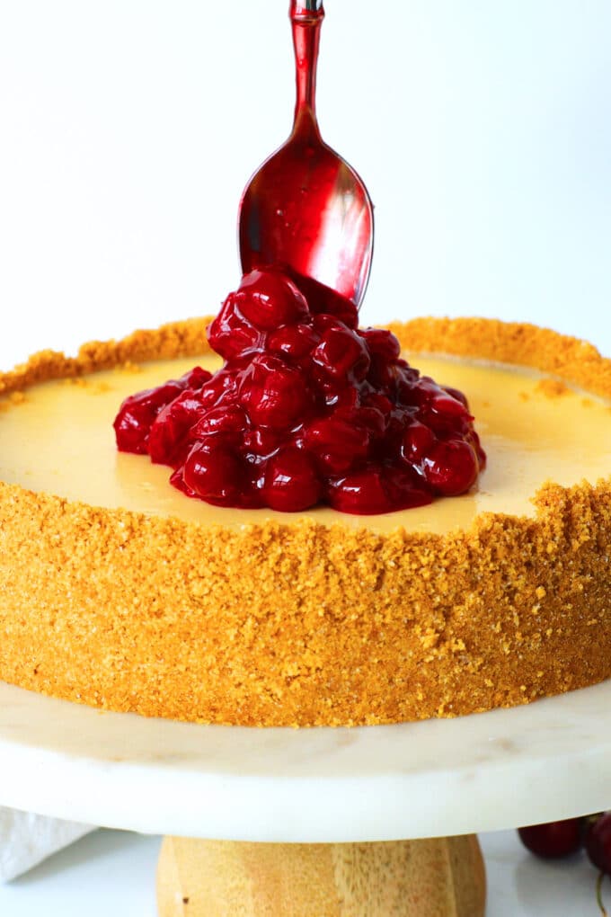 cherry topping being spooned onto the top of the cherry cheesecake sitting on a cake plate