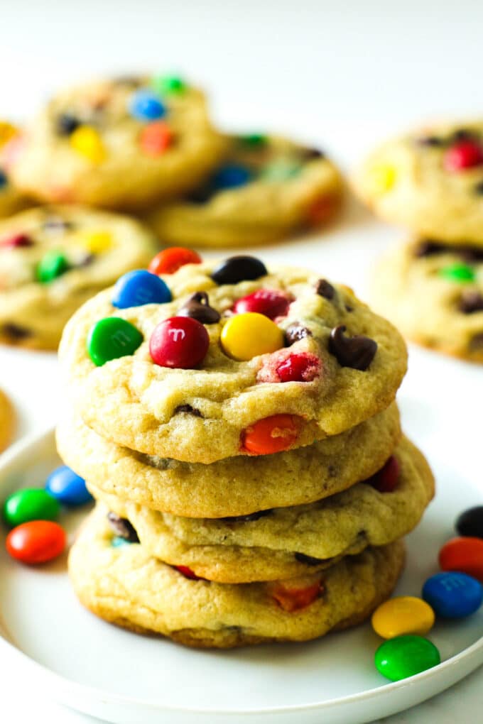 A stack of M&M cookies sit on a plate with M&Ms beside them and other cookies in the background. 