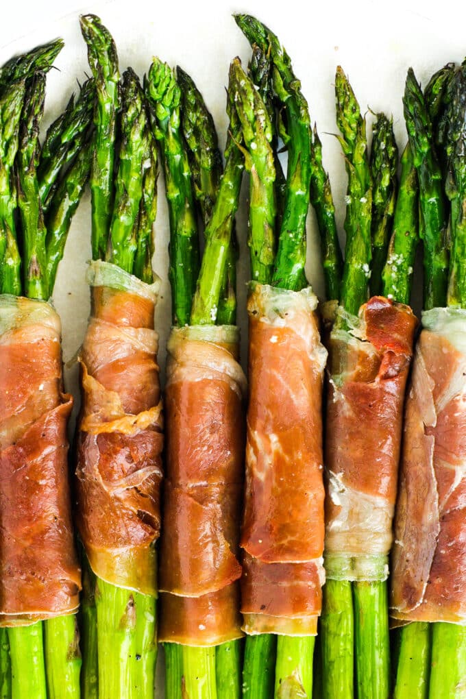 A close up of prosciutto wrapped asparagus bundles laying close to each other on a white plate