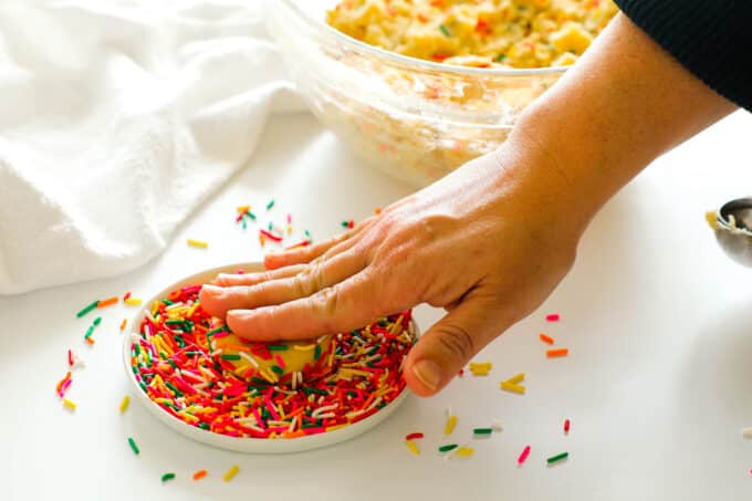 A hand pressing a round of cookie dough into a plate of sprinkles
