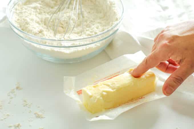 a persons finger making an indention in an unwrapped stick of butter, a bowl of flour in the background