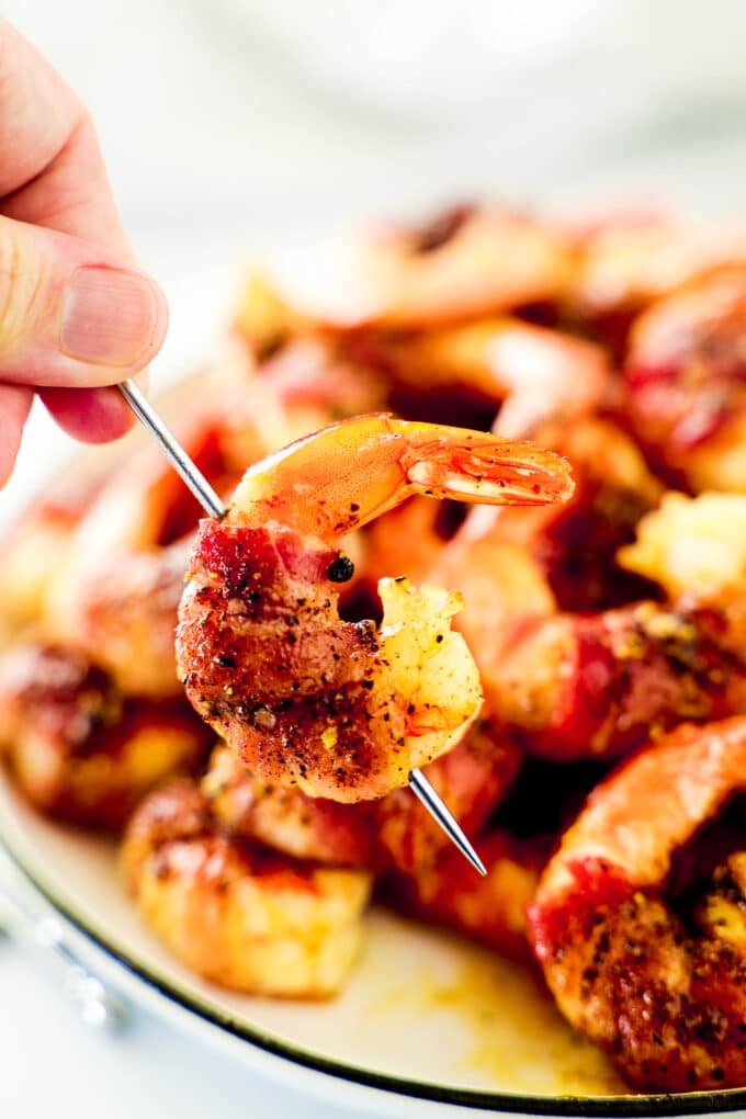 A hand holding a Bacon Wrapped Shrimp on a metal pick. The rest are on a plate in the background.