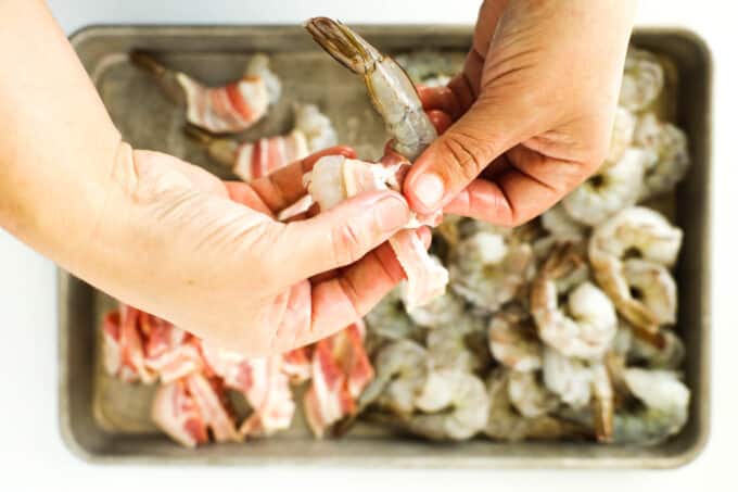 Hands wrapping half a piece of bacon around a single raw shrimp.