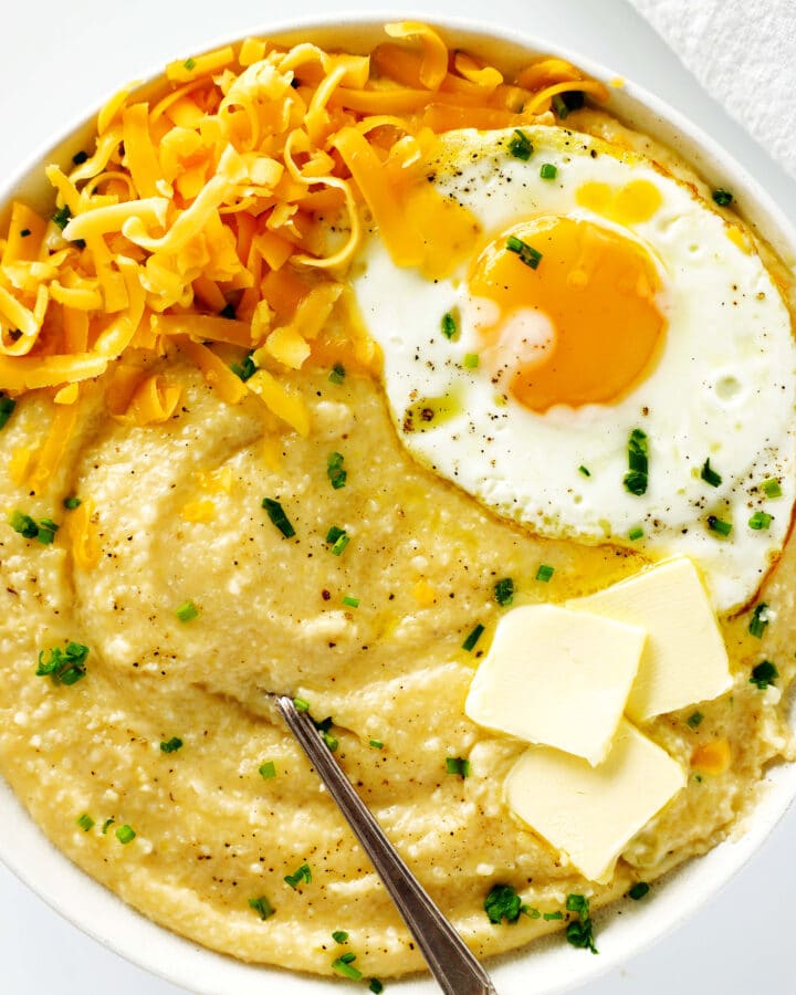 An overhead view of a bowl of cheese grits with a fried egg, shredded cheddar, pats of butter, and chopped chives on top.