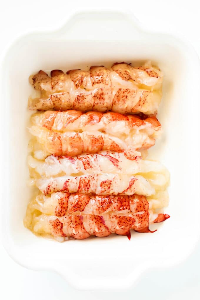 Four lobster tails with the shells removed laying in a row in a baking dish.