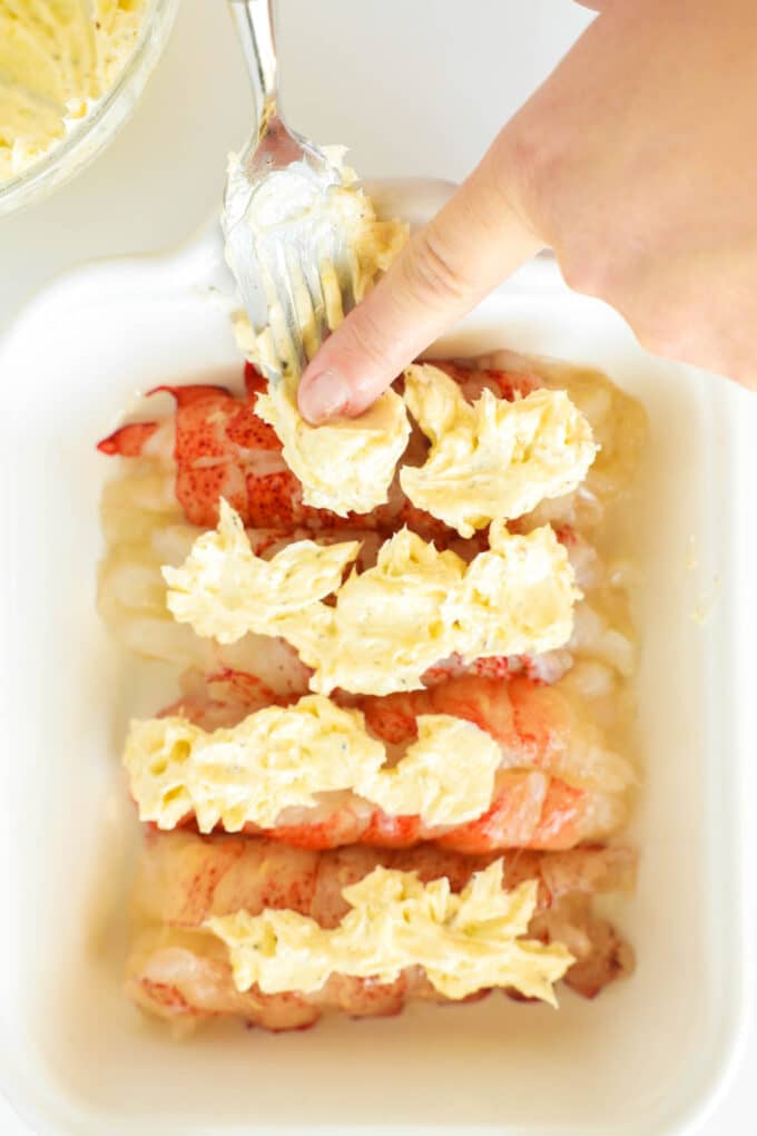 A hand pushing scoops of the garlic butter mixture from a fork onto the lobster tails in the baking dish.