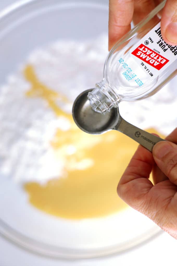Peppermint extract being measured out over a bowl of powdered sugar and sweetened, condensed milk.
