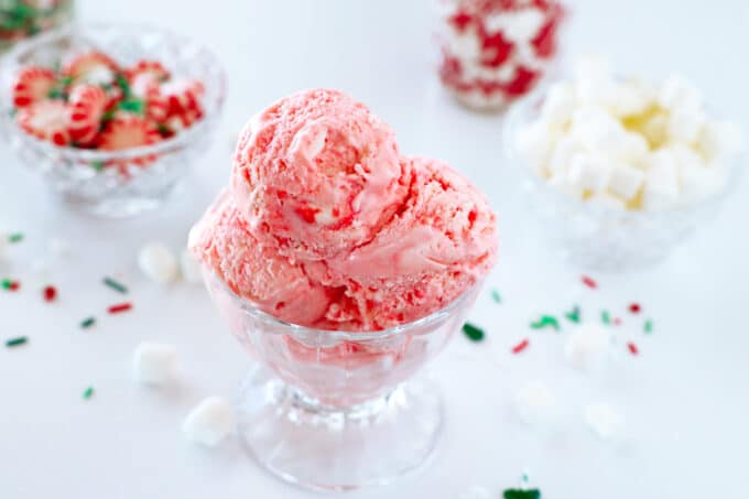 A glass ice cream dish heaping full of pink Peppermint Ice Cream sits on the counter with bowls of peppermint candies and mini marshmallows behind it.