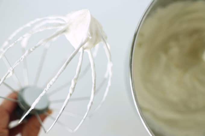 The whisk attachment of a stand mixer being held up to show the stiff peak on the end of it.